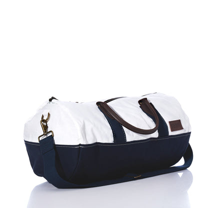 The Chebeague Duffel by Sea Bags is crafted from recycled sail in white and navy blue fabric, featuring premium leather handles, a detachable navy shoulder strap, and a brown patch on the side, shown on a white background. by Sea Bags