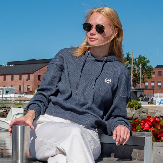 A woman wearing the Soft As A Grape Ribbed Hoodie Pullover - Navy Anchor and sunglasses sits outdoors by a harbor, holding a metal tumbler, with boats, buildings, and flowers visible in the background.