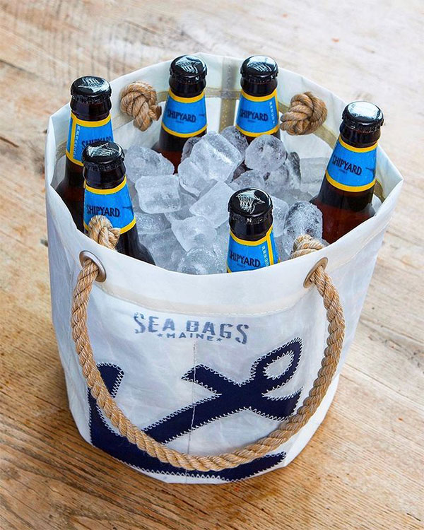 a white recycled sail cloth beverage bucket with hemp rope handles, the Sea bags logo, and a navy anchor, sits on a table. The bucket is filled with six glass bottles and ice.