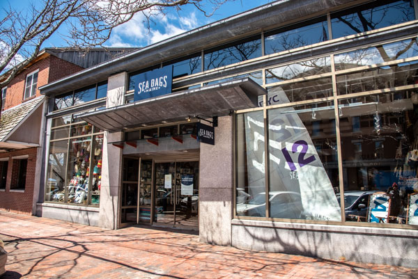 front view of a Sea Bags store, double doors in the middle and large windows on either side. In the left window is a scattering of bouys attached to a fishing net. In the right window is a large white sail with a purple 12 insignia.