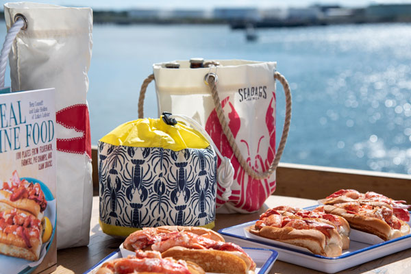 with the ocean in the background, a restaurant table is covered with recycled sail cloth bags featuring lobster prints and two trays of lobster rolls in the foreground