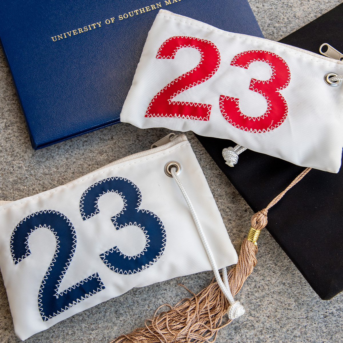 two white recycled sail cloth wristlets with 23 insignia sit on a granite step with a grad hat and diploma