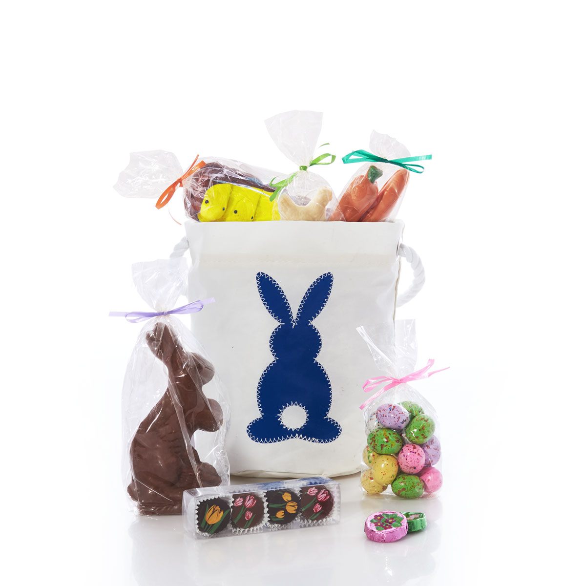 a blue silhouette of bunny with a white circle tail on a white recycled sail cloth bucket bag with a white rope handle is filled with packaged Easter themed chocolate and candy treats