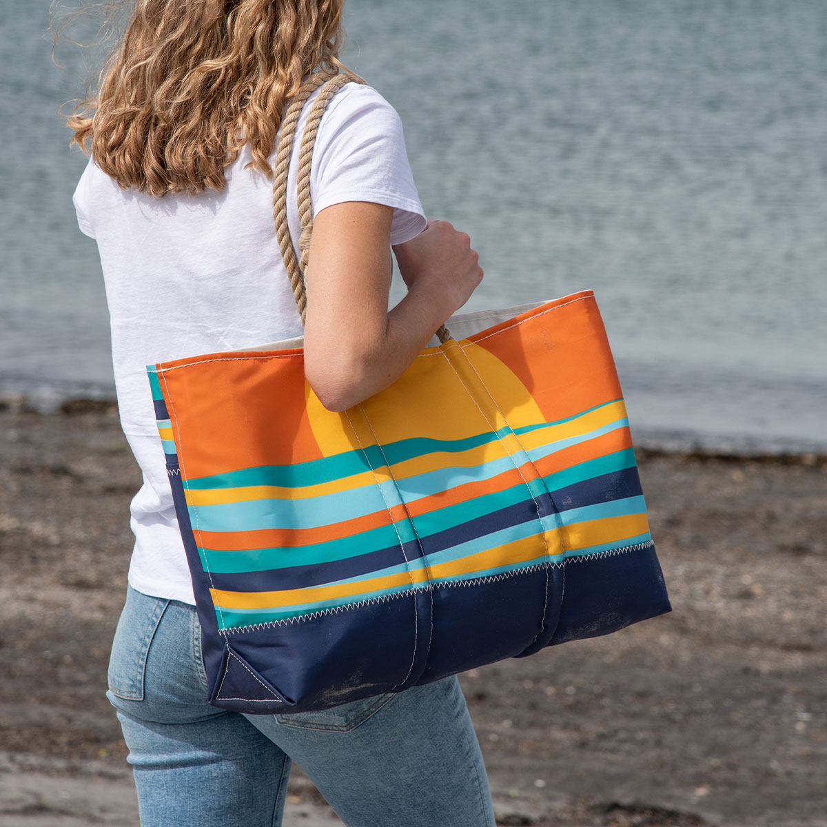 a person is holding a tote with multicolor wavy ocean stripes sit under a rising sun printed on a recycled sail cloth tote with hemp rope handles