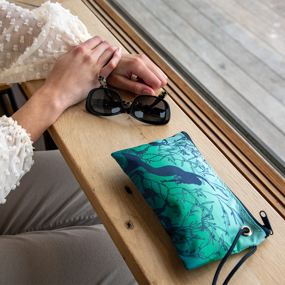 a mermaid print wristlet sits on a counter next to a pair of sunglasses and a woman with crossed hands