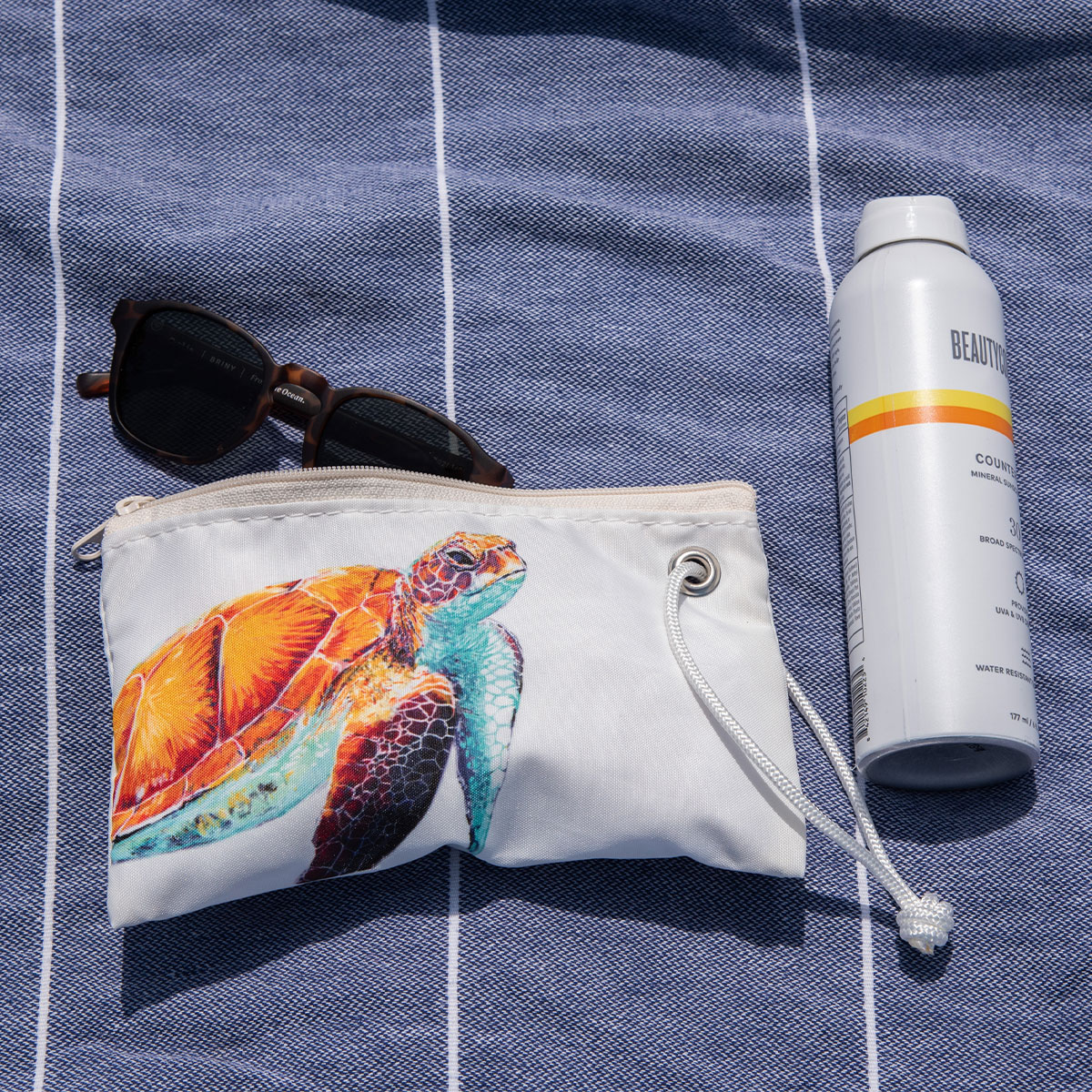 a bright friendly multicolor sea turtle is printed on a white recycled sail cloth wristlet sits on a blue beach towel next to a water bottle