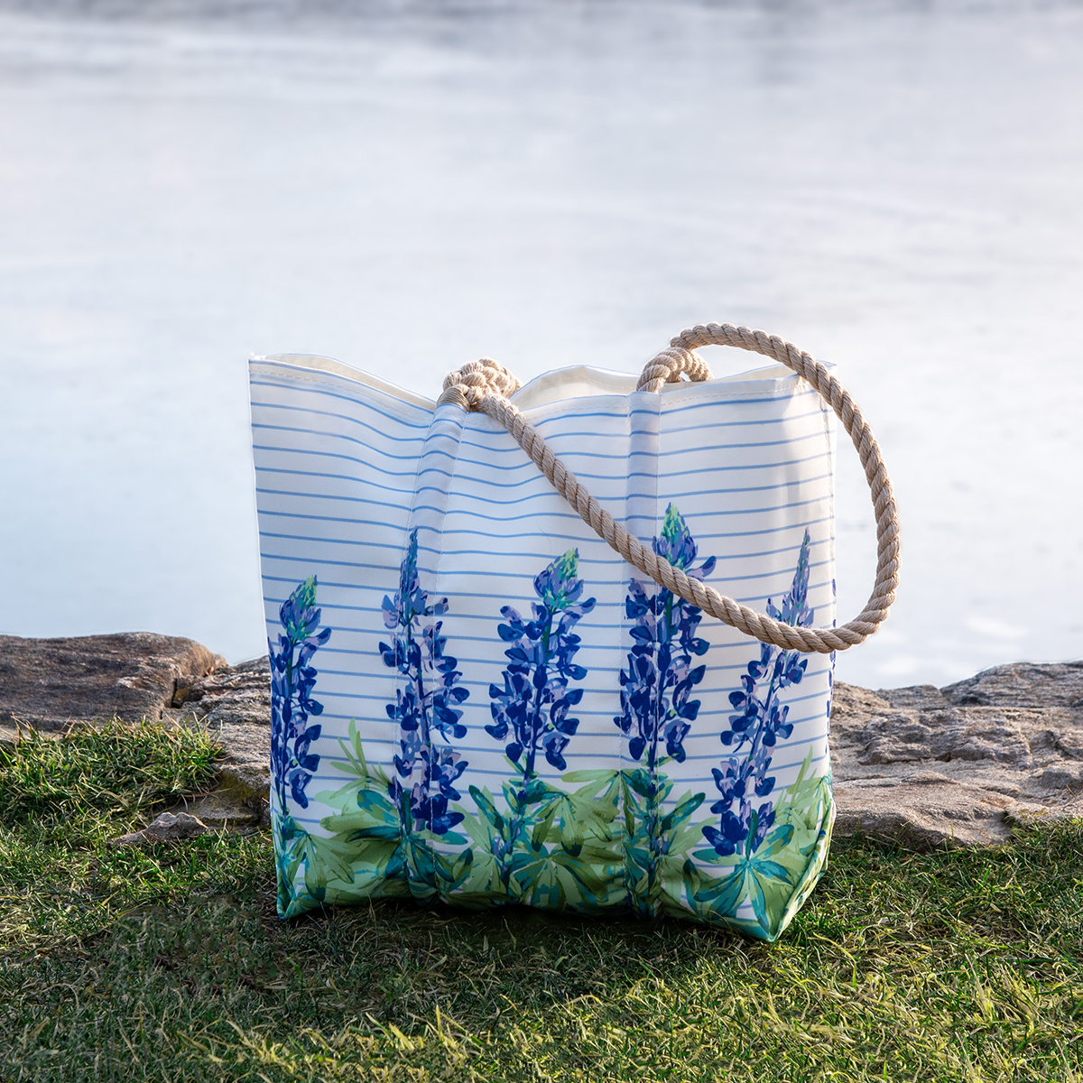 a recycled sail cloth bucket bag is printed with thin blue stripes behind a row of purple and green lupine flowers and finished with a hemp rope handle sits on grass by the water