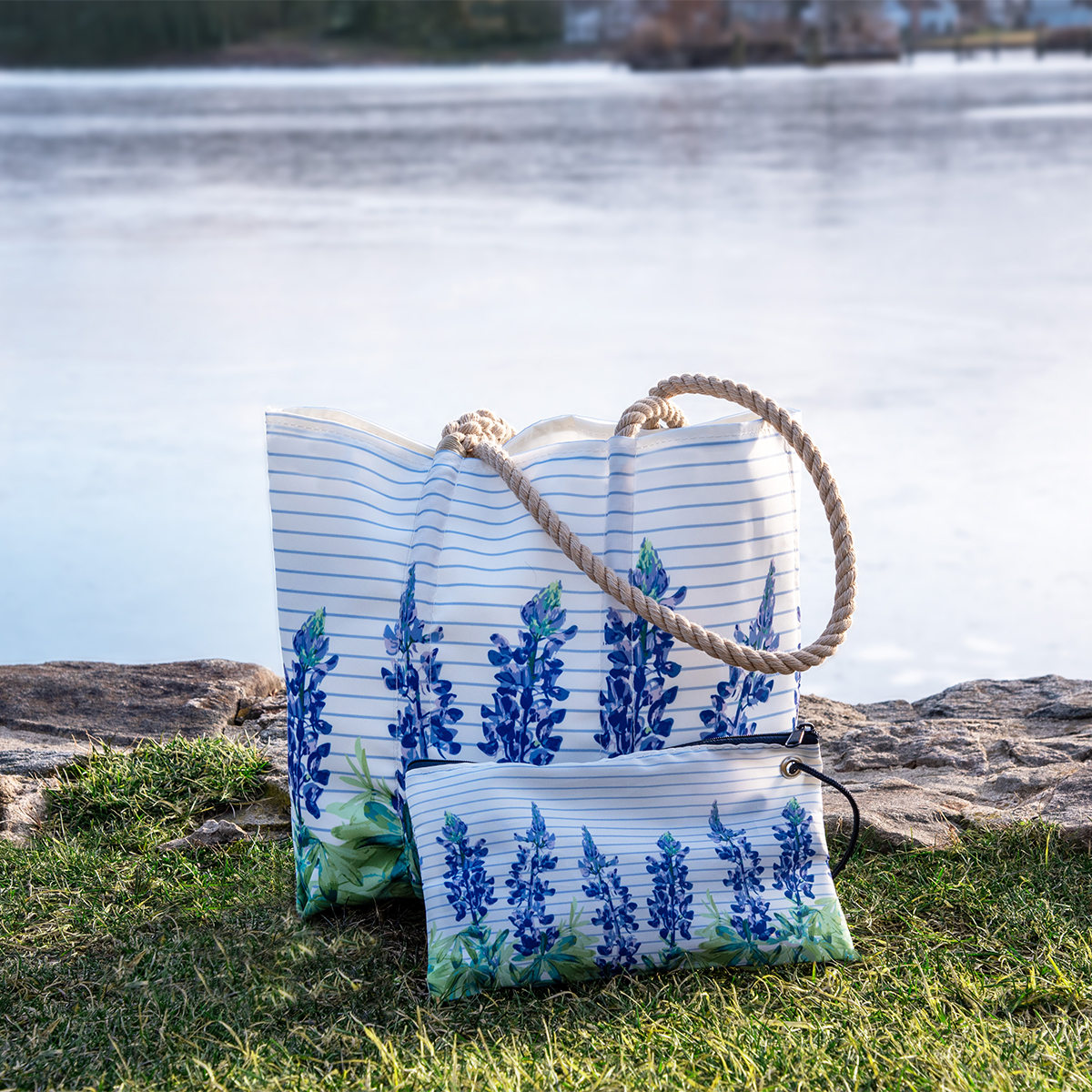 a lupine sailor stripe large wristlet sits with a lupine sailor stripe medium tote on a grass patch by the ocean
