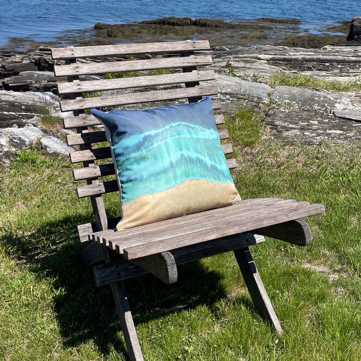 sitting on an outdoor chair is a recycled sail cloth pillow is printed with stripes of tan, teal, and blue depicting waves washing onshore