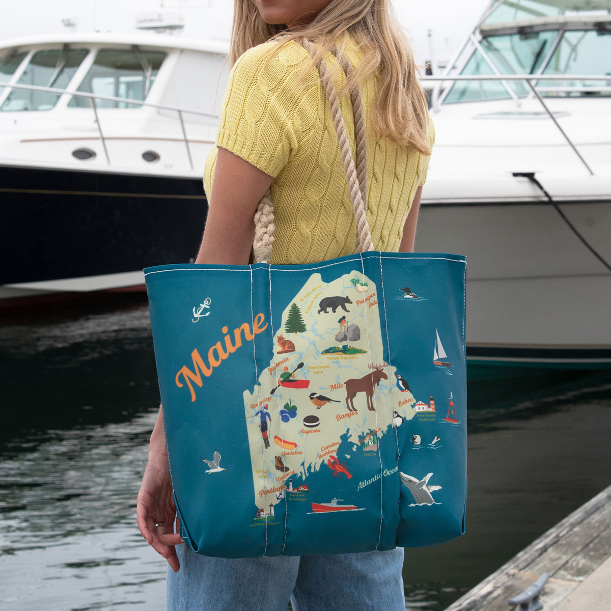 a person is wearing a tote showing a green state of Maine is printed on a blue recycled sail cloth tote with hemp handles and is filled with icons depicting unique landmarks