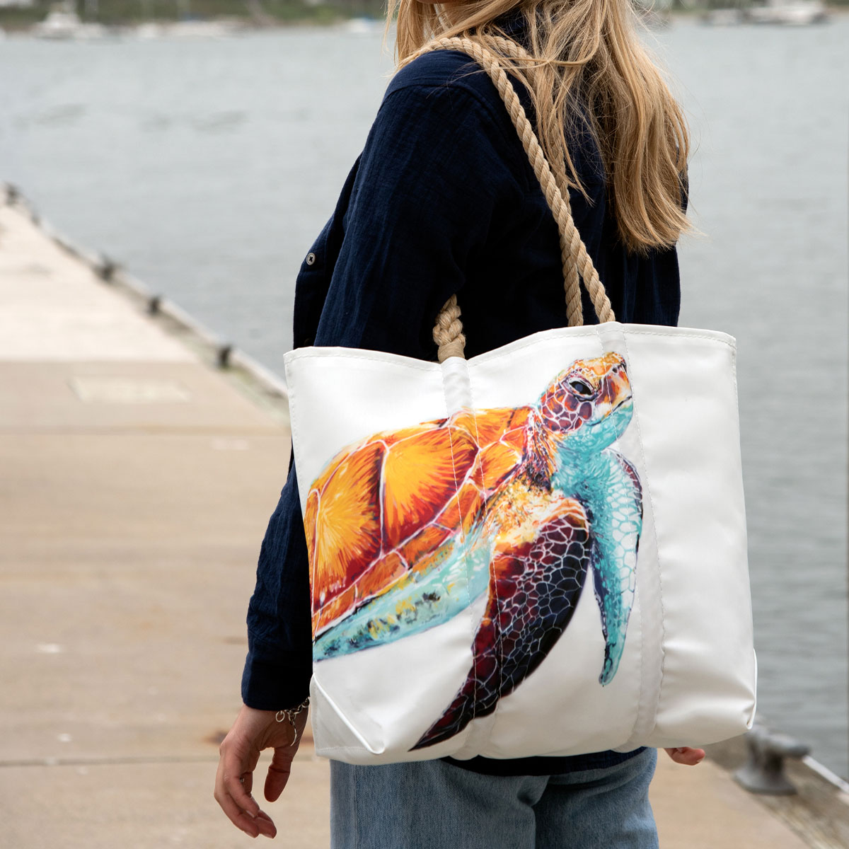 a person is wearing a tote with a bright friendly multicolor sea turtle is printed on a white recycled sail cloth tote with hemp rope handles