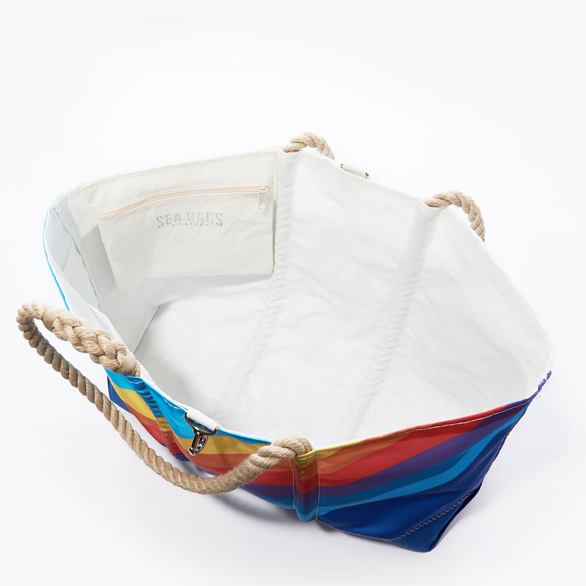 inside view showing hanging interior zipper pocket of a recycled sail cloth ogunquit beach tote which is printed with retro stripes that range from light blue in the top left corner to dark blue in the bottom right corner, with rainbow colored stripes in between