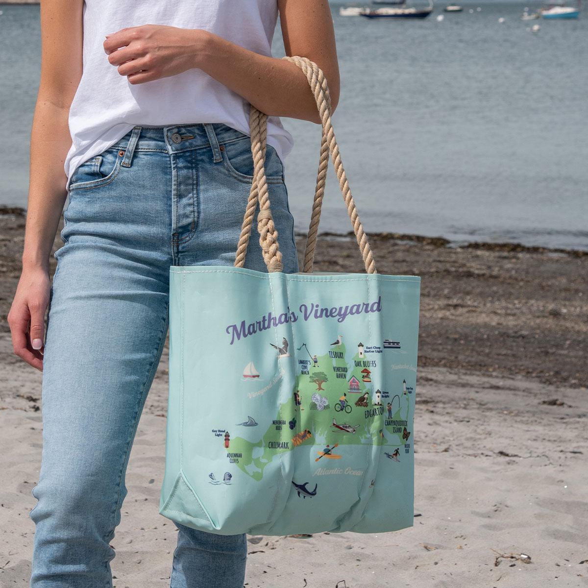 a person is wearing a tote showing a green area of Martha's Vineyard is printed on a blue recycled sail cloth tote with hemp handles and is filled with icons depicting unique landmarks