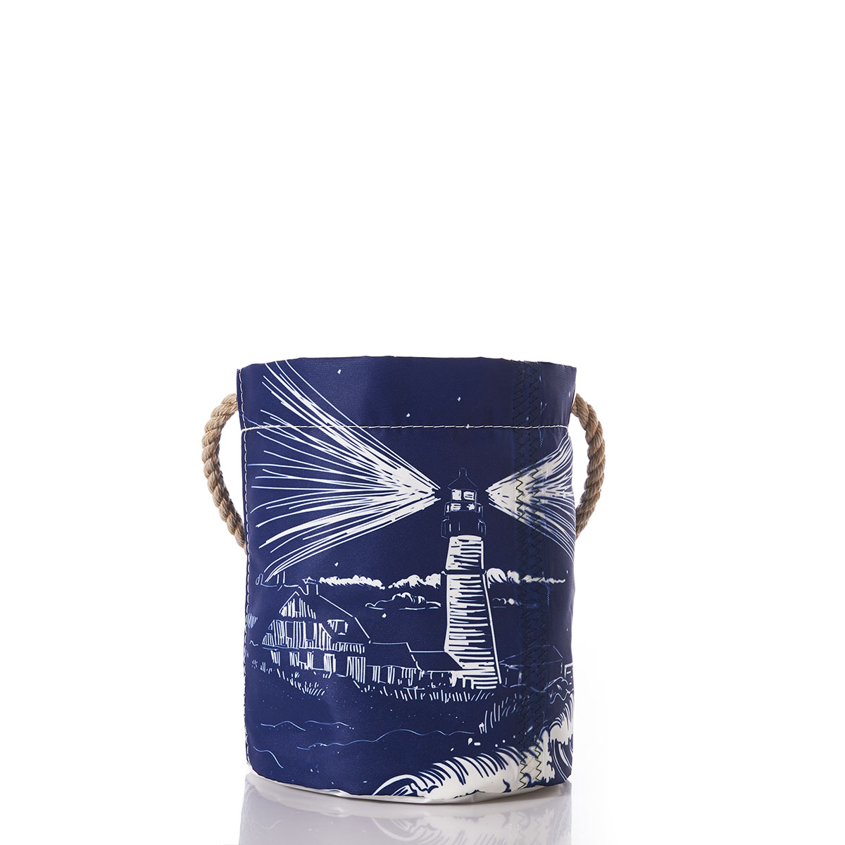 a recycled sail cloth bucket with hemp rope handles is printed with a navy background and a white image of a lighthouse on a coastal rock with waves crashing and the light beaming through the dark